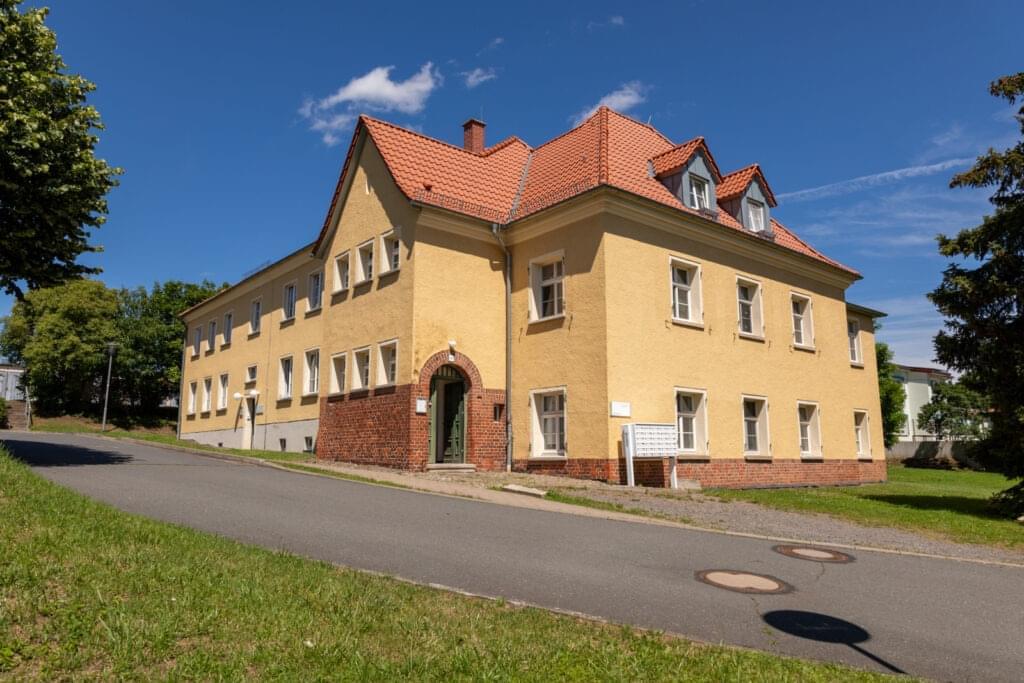 An older, yellow building with a red tiled roof. The sky is blue. A narrow road runs in front of it.