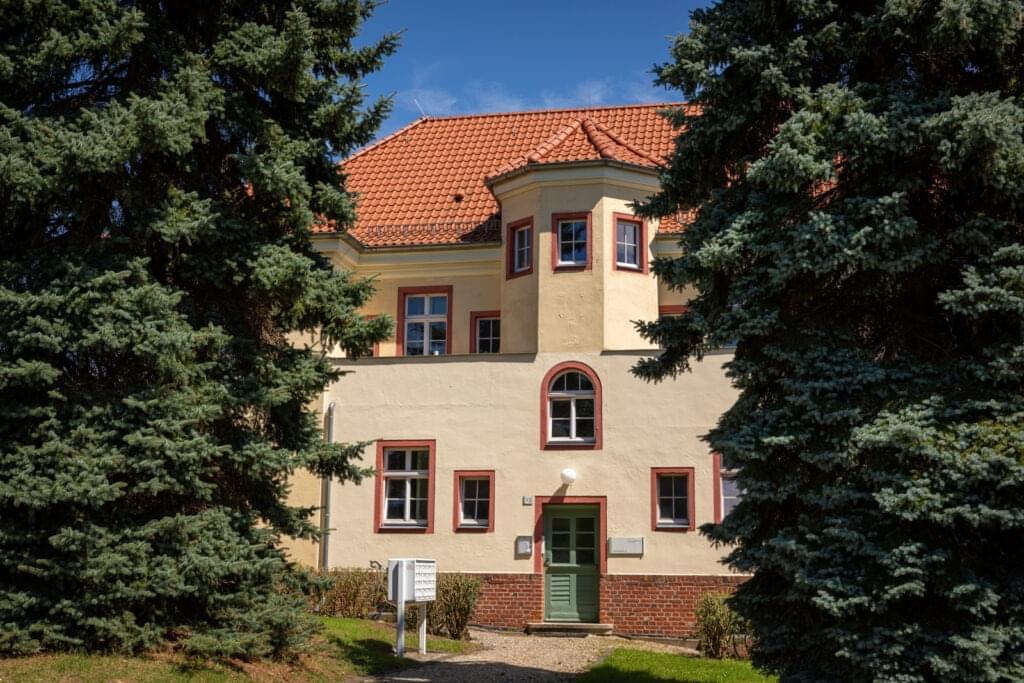 An older building with a red tiled roof and a small tower. There are conifers to the left and right.