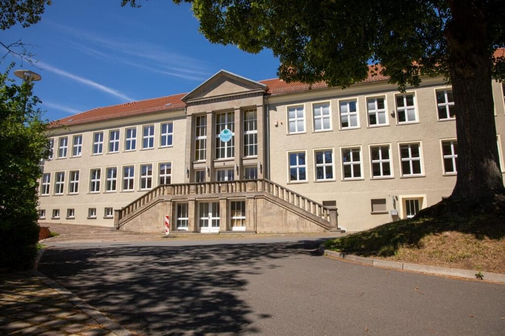Wohnheim des Studienkollegs auf dem Campus Blick von leicht schräg unten auf die Vorderseite des Gebäudes 12 - Mittag der Eingangsbereich mit der Freitreppe,, rechts steht ein Baum. Der Himmel ist blau.